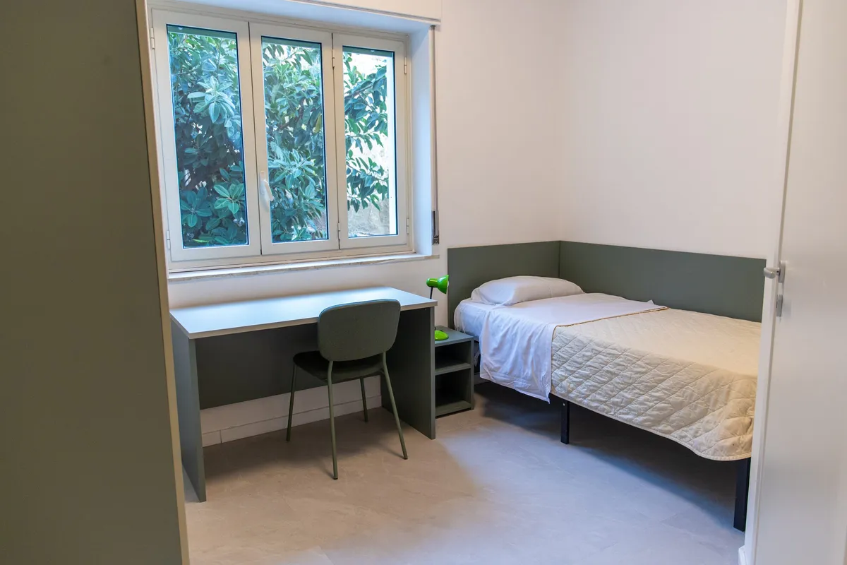 Habitación individual con escritorio, silla, mesita y cama bajo una ventana con vista a los árboles